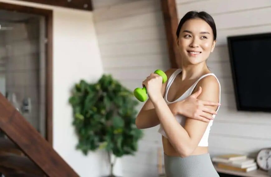 A smiling woman exercising with a green dumbbell at home.