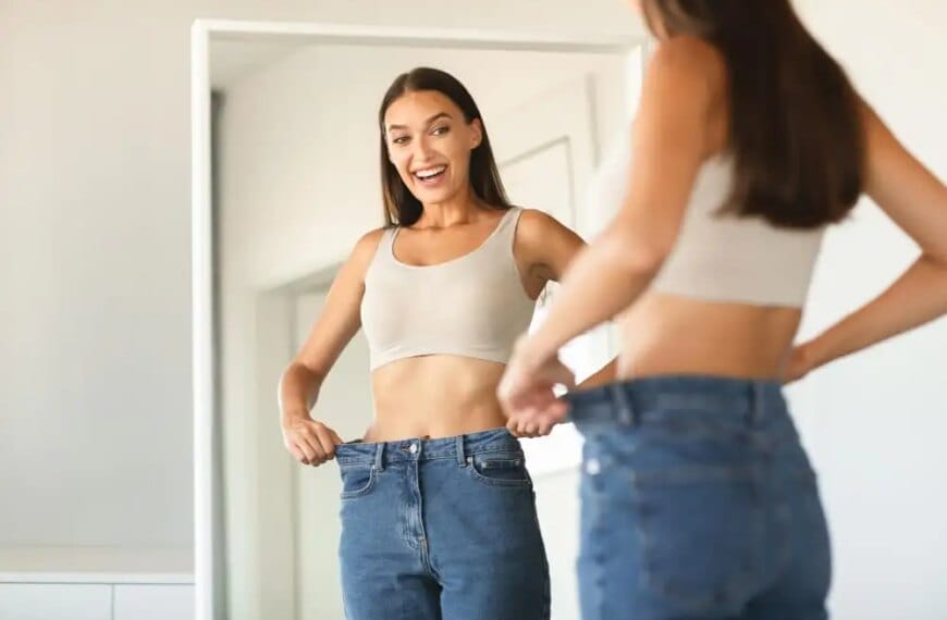 Woman smiling at mirror, holding oversized jeans, showcasing weight loss.